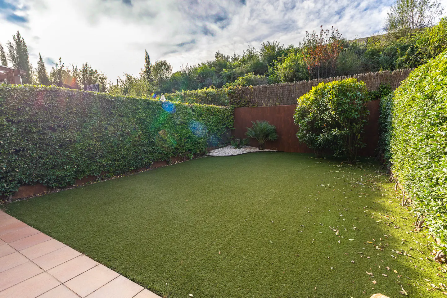 A backyard with green artificial turf, surrounded by tall green hedges and a brown fence.