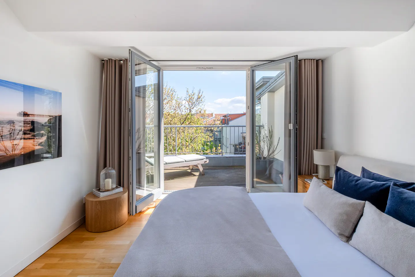Modern bedroom with a queen bed, beige bedding, and open glass doors leading to a sunny balcony with a wooden deck.