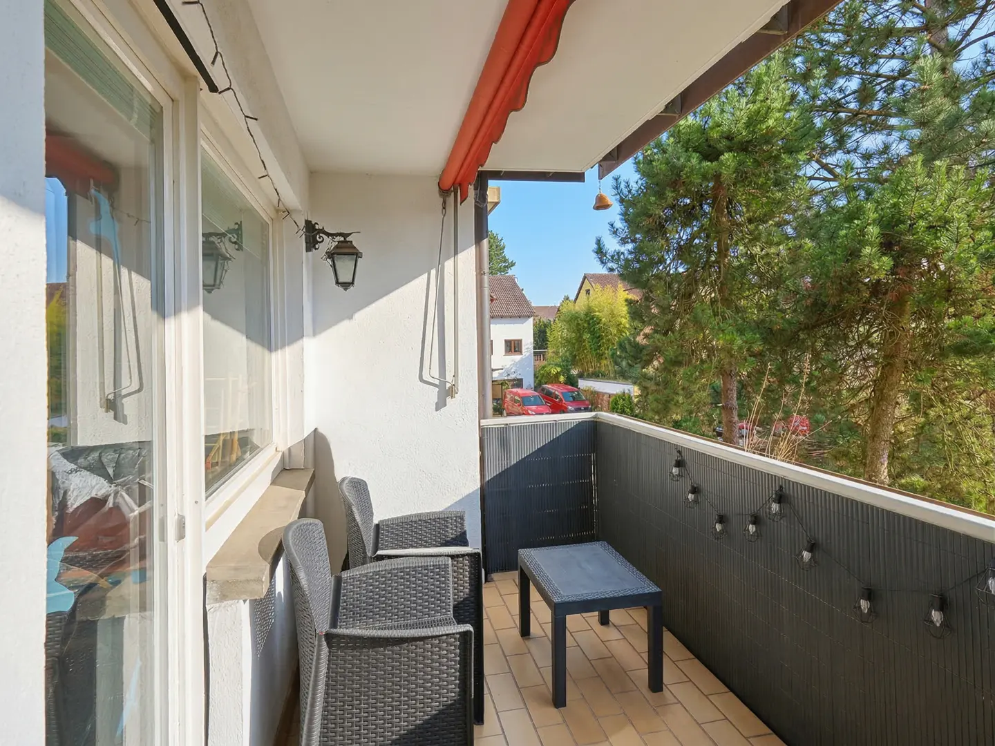 A balcony with a table and chairs overlooks trees and houses. The balcony has a red awning and string lights.