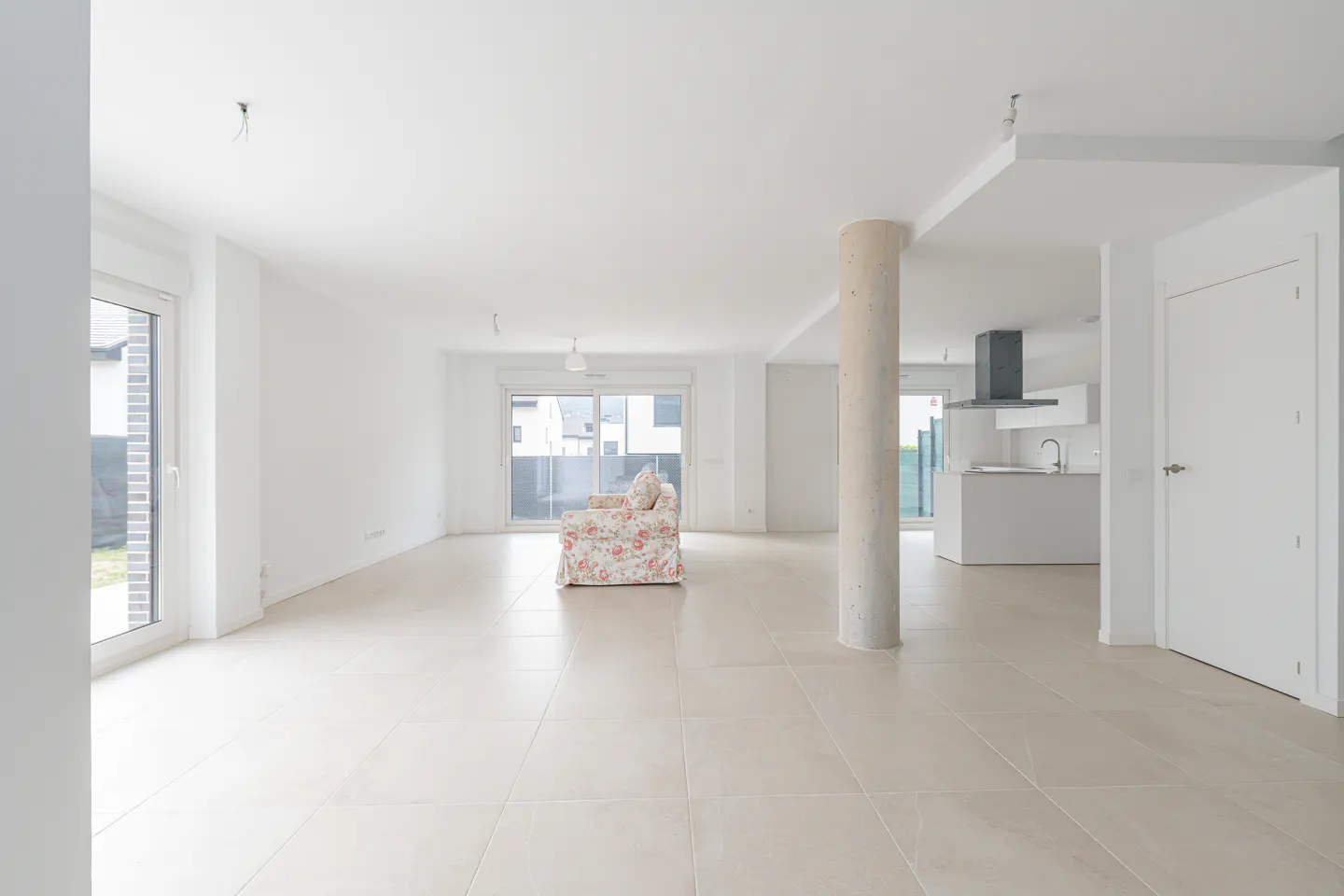 Bright, open-plan living space with white walls, tiled floor, and a floral sofa. A concrete pillar separates the living area from the kitchen.