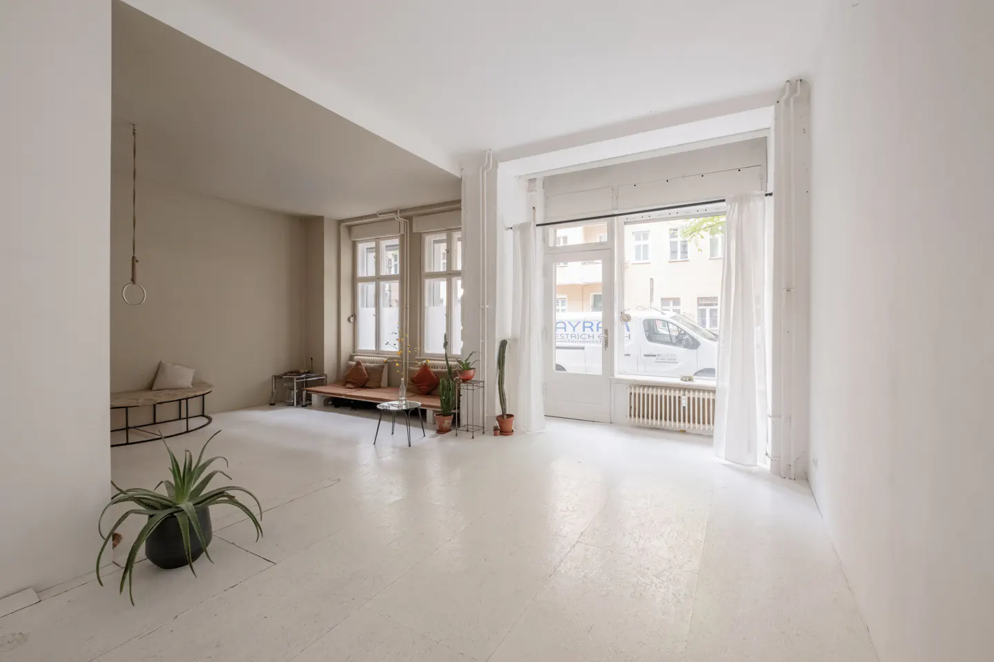 Bright, airy room with white floors, neutral walls, and large windows. Plants, benches, and a glimpse of a street scene outside.