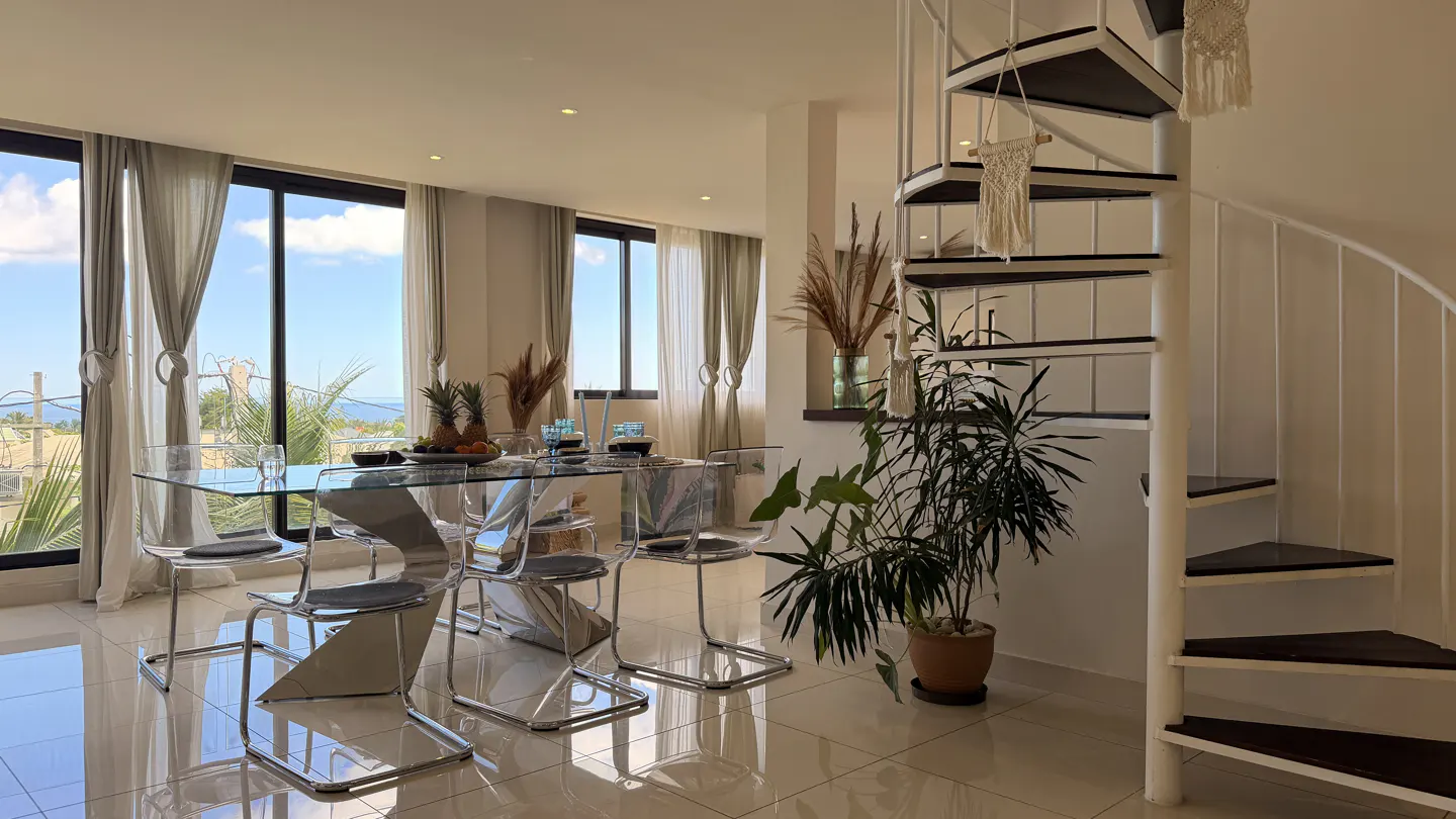 Bright dining room with a glass table, clear chairs, and a spiral staircase. Large windows offer a view of the sky and ocean.