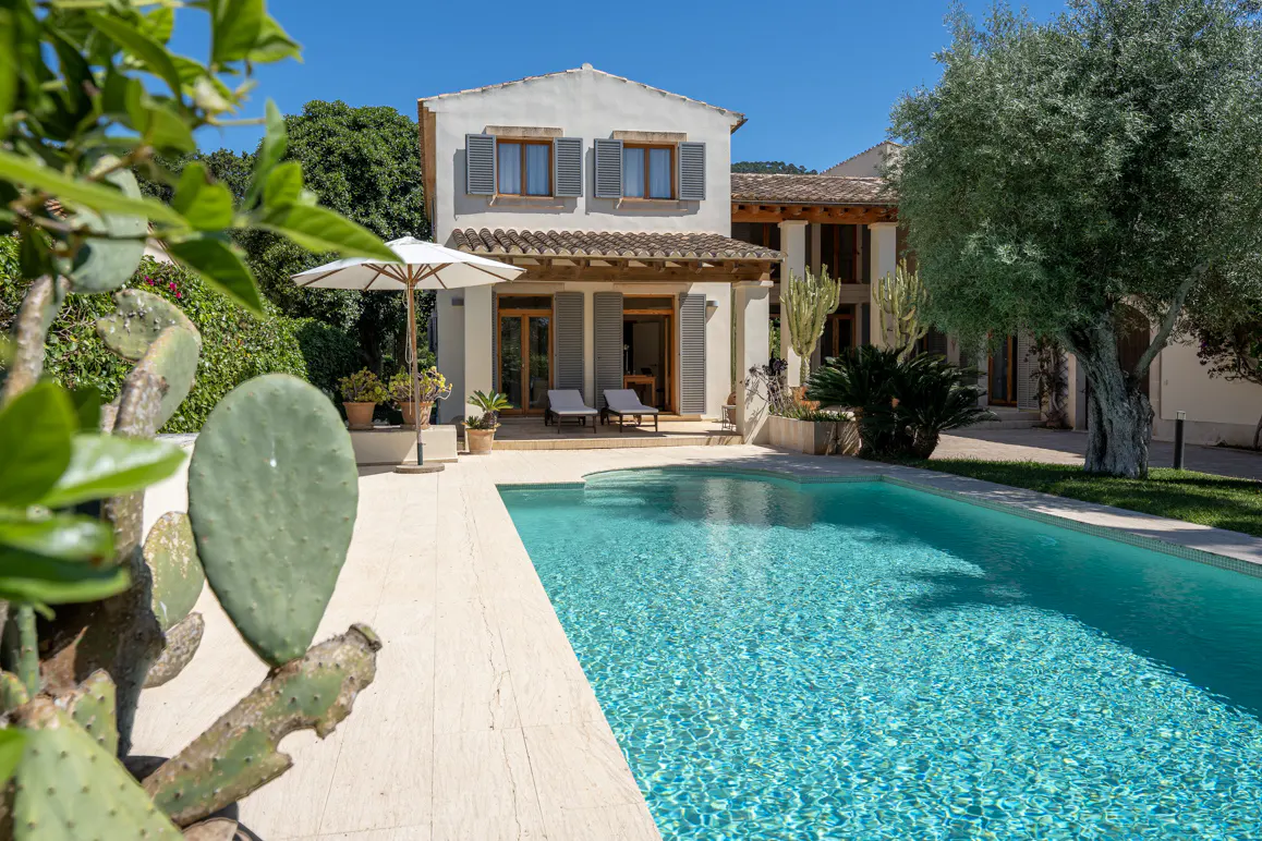 A Mediterranean-style villa with a swimming pool, sun loungers, and a white umbrella, framed by cacti and olive trees.
