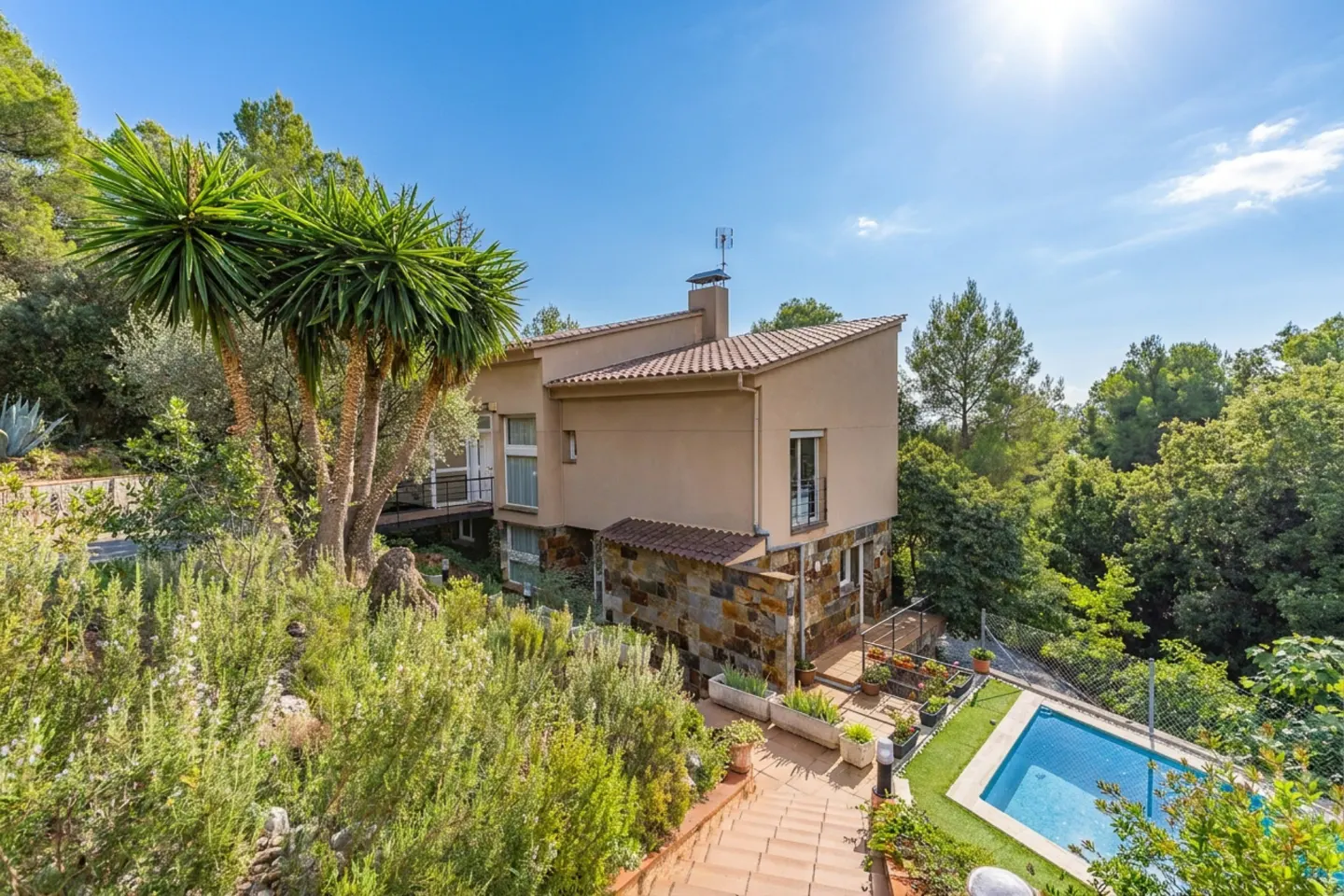 Exterior view of a modern beige house with a stone facade, surrounded by lush greenery and a blue swimming pool.