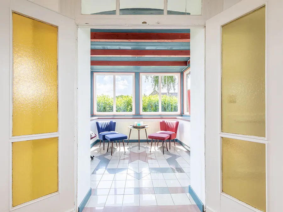 View through a doorway into a sunroom with blue and red chairs, a small table, and a geometric tiled floor.