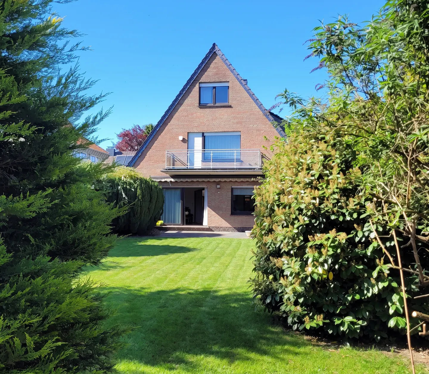 Backyard view of a two-story brick house with a balcony, green lawn, and trees under a clear blue sky.