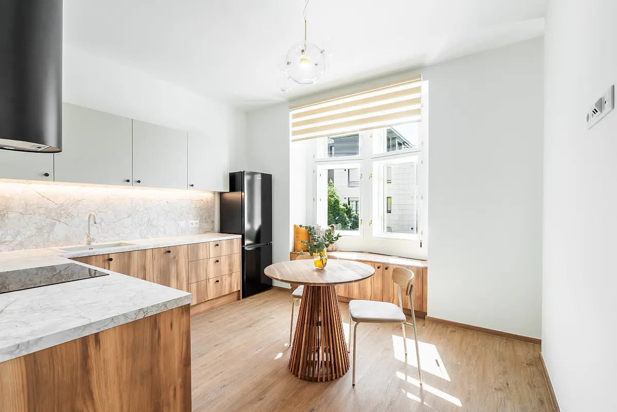 Bright kitchen with wood cabinets, marble counters, and a black fridge. A round table sits near a window with a built-in bench.