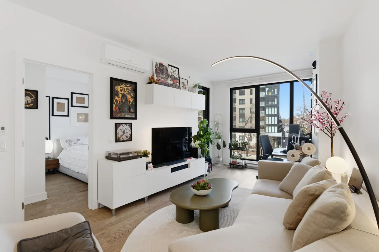Bright living room with white walls, a beige sofa, and a green coffee table. A doorway leads to a bedroom with a white bed. Large windows offer city views.