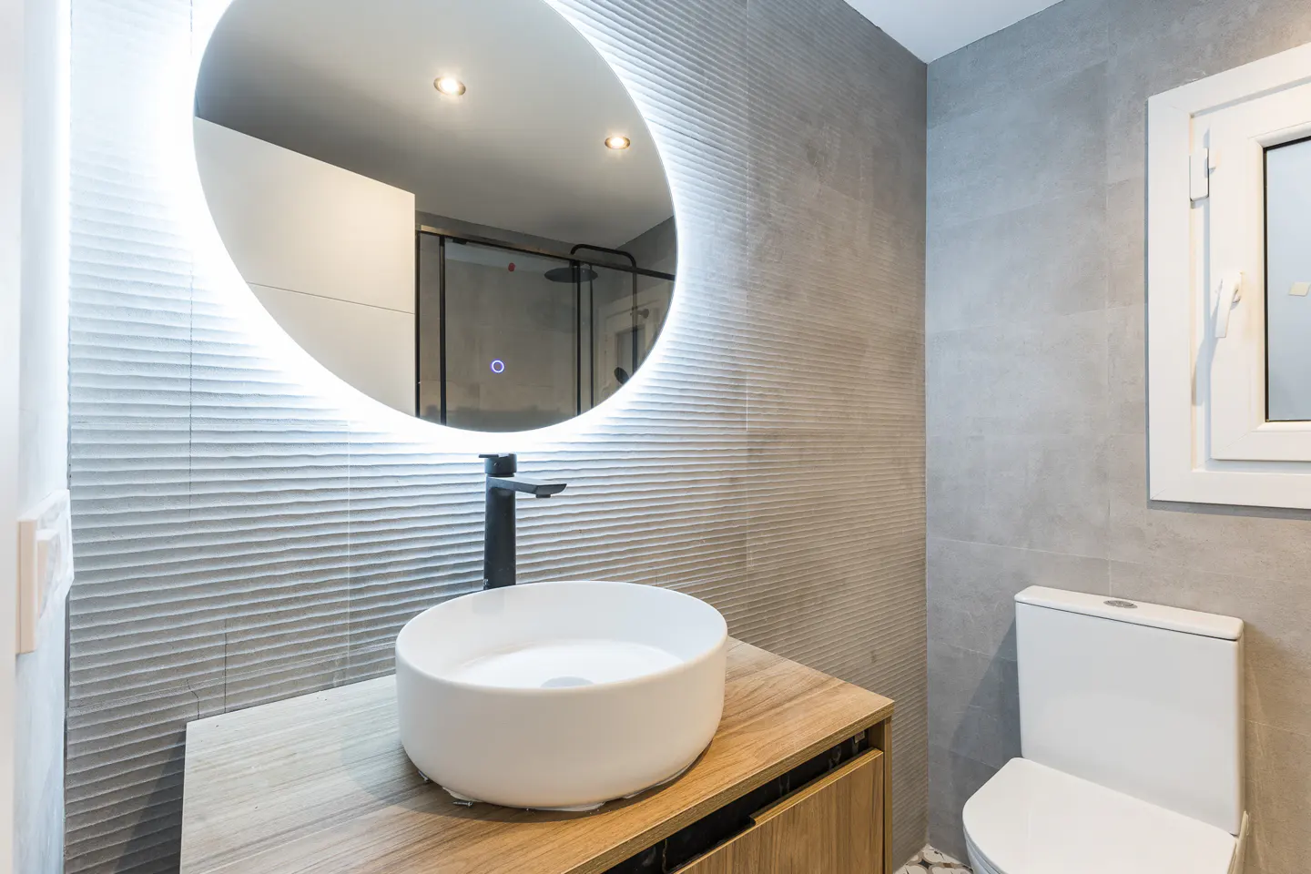 Modern bathroom with gray textured walls, a round LED mirror, a white vessel sink on a wood vanity, and a white toilet.