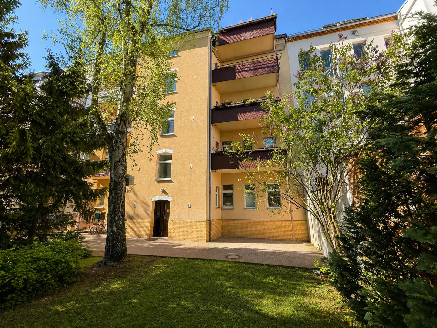 Exterior view of a yellow apartment building with brown balconies, surrounded by green trees and grass.