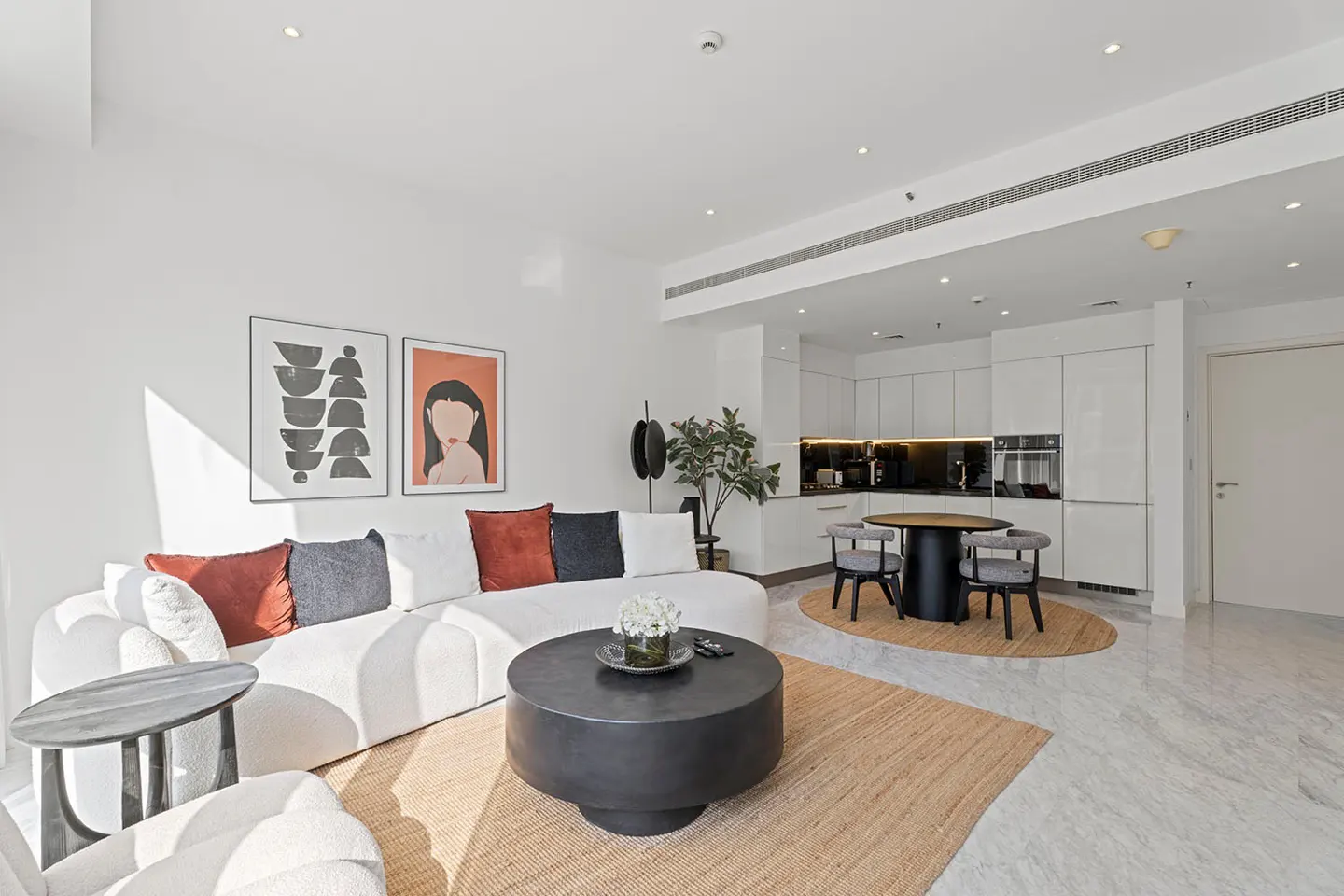 Bright, modern apartment interior with a white sofa, black coffee table, and open kitchen with white cabinets. Artwork adorns the walls.