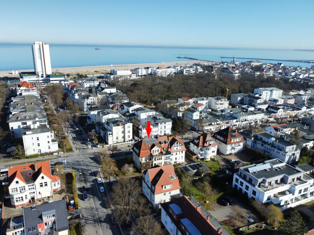 Aerial view of a coastal town with a red arrow pointing to a specific multi-story building with a red roof.