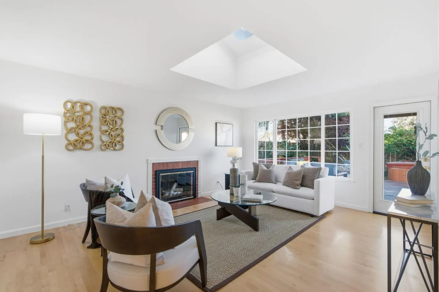 Bright living room with white walls, wood floors, and a skylight. A white sofa faces a fireplace, with chairs, a rug, and a glass table in between.