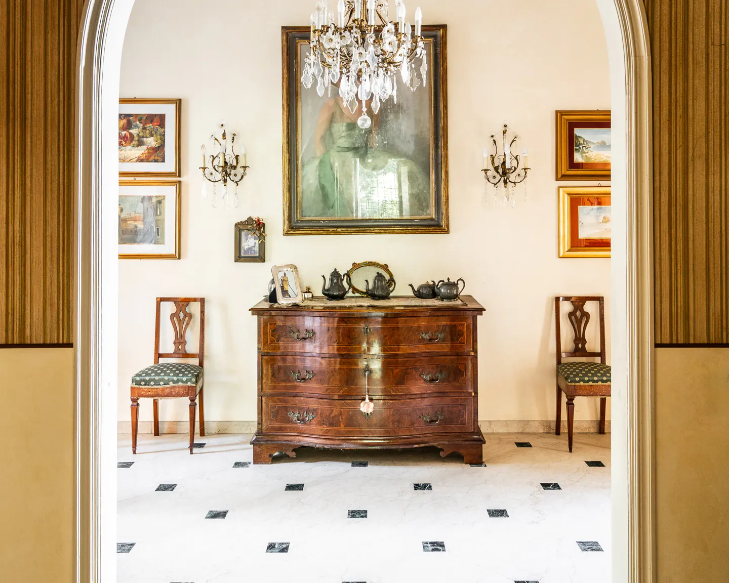Interior view through an archway of a room with a dark wood dresser, chairs, and framed art on the walls. A chandelier hangs above the dresser.