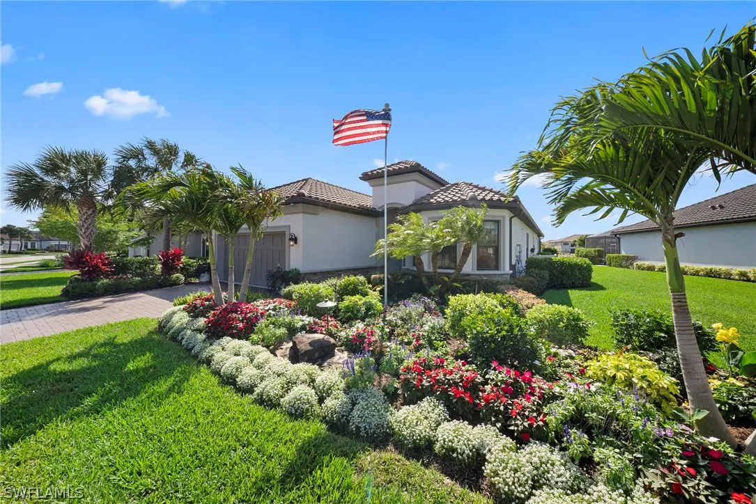 Exterior of a one-story house with a tile roof, palm trees, and a lush garden. An American flag waves in front of the house under a blue sky.