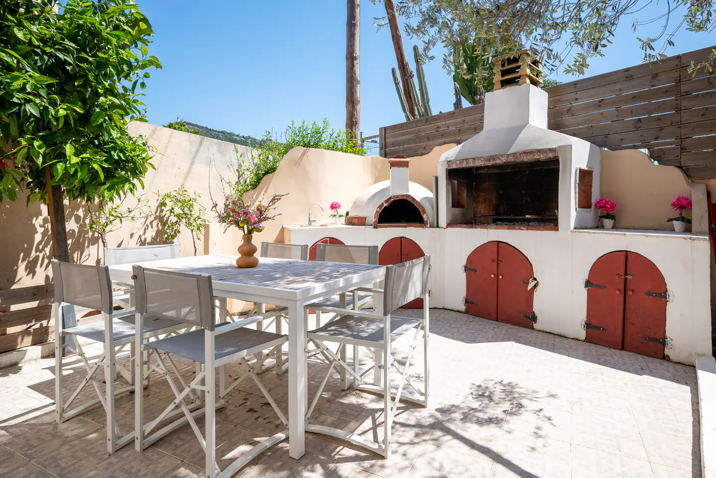 Outdoor dining area with white table and chairs, a built-in grill, pizza oven, and red doors on a sunny patio.