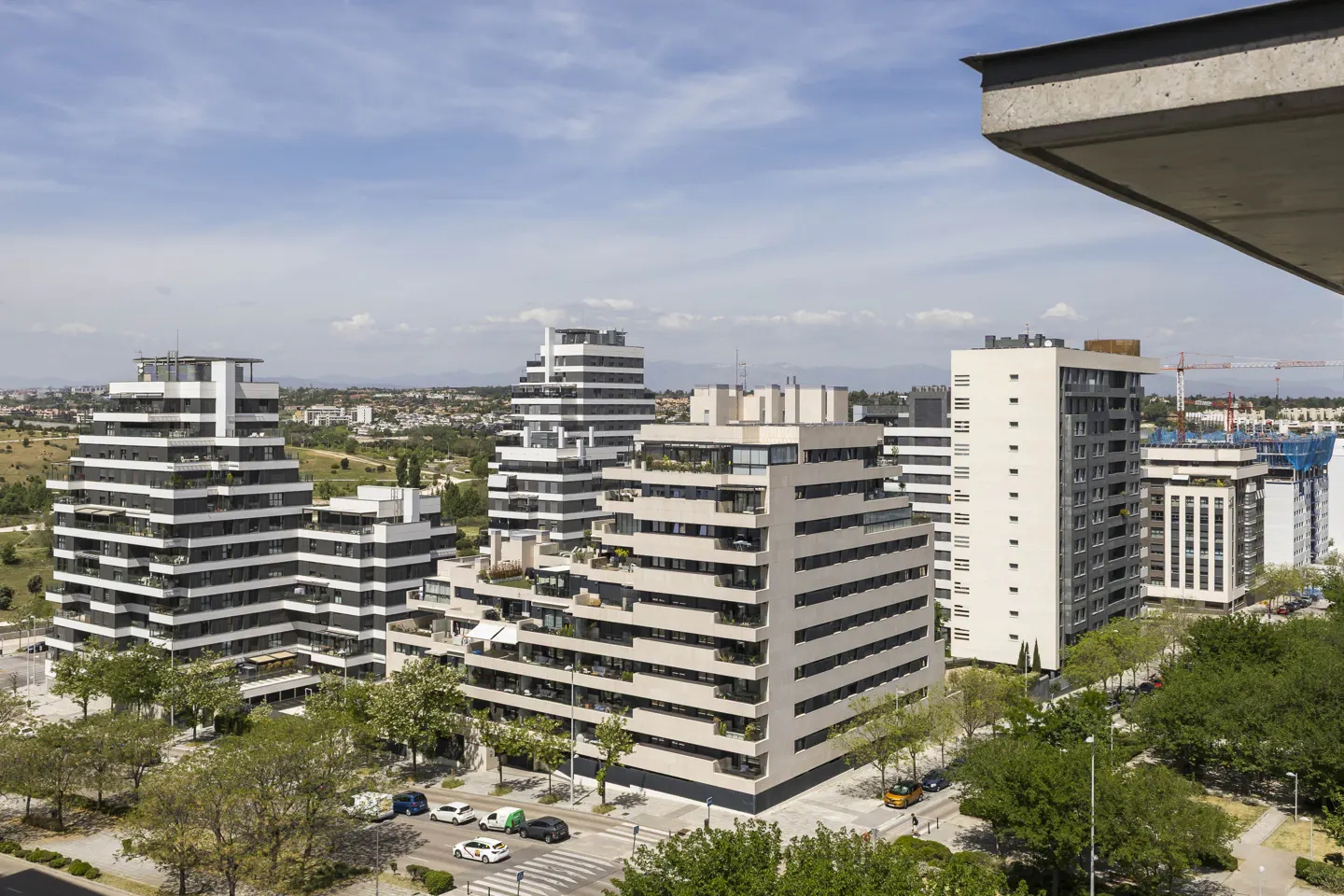 View of modern, tiered white apartment buildings with balconies, trees, and cars on a city street under a blue sky.