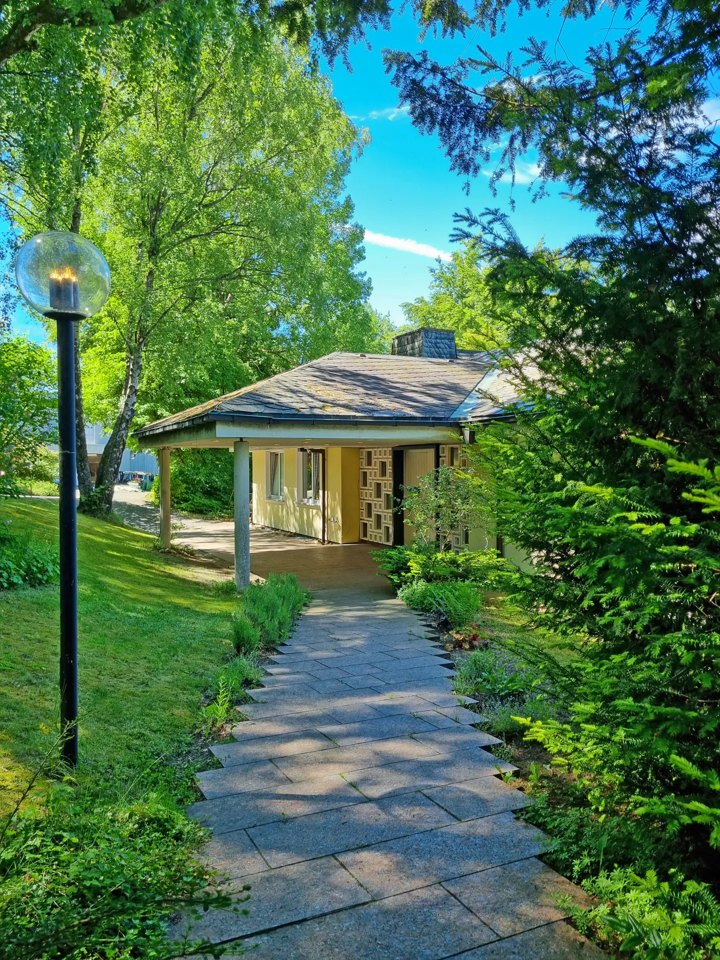 Exterior view of a yellow house with a covered entrance, stone walkway, and lush green landscaping under a blue sky.