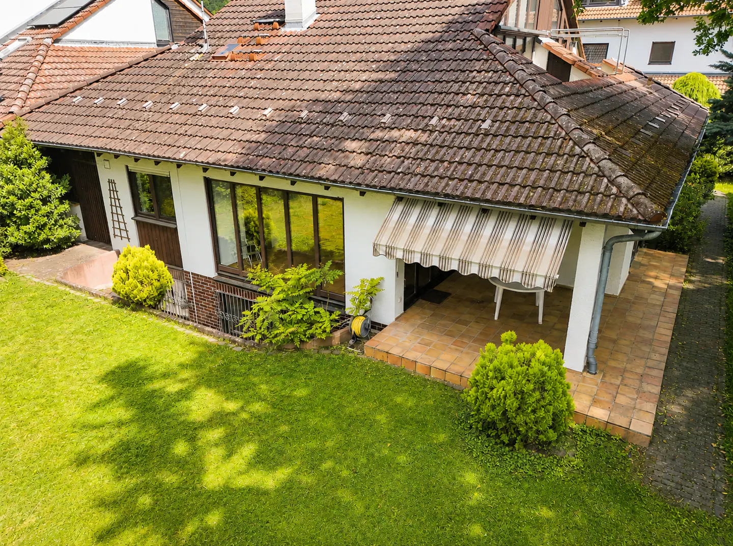 Aerial view of a one-story house with a brown tiled roof, white walls, large windows, and a patio with a striped awning. Green lawn and trees surround the house.