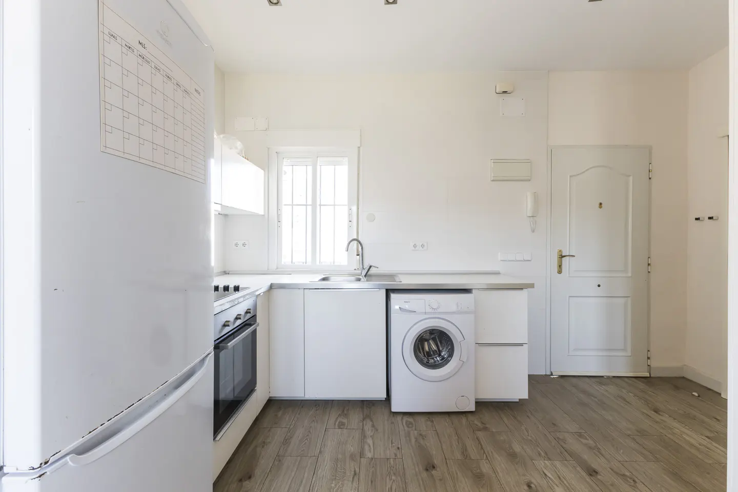 Bright, all-white kitchen with wood-look floors, a fridge with a calendar, oven, sink, and a washing machine.