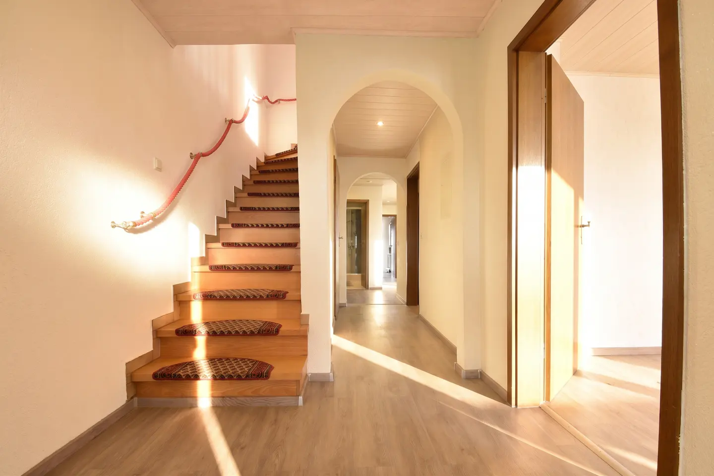 Interior view of a home featuring a staircase with carpet treads, a red rope handrail, and an arched hallway with open doors.