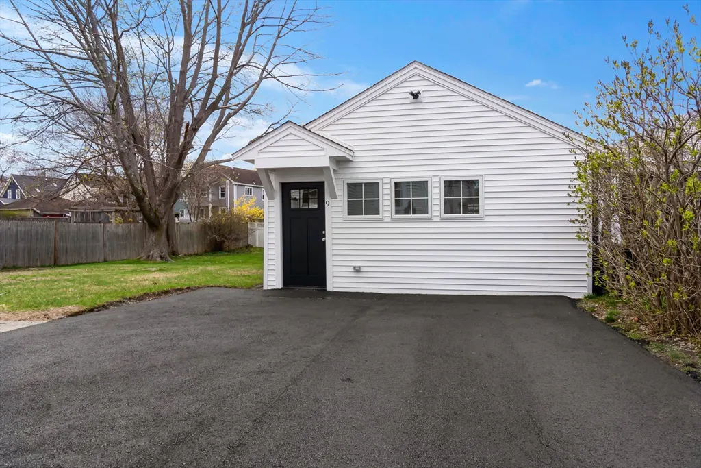 Exterior view of a white, one-story house with a black door and asphalt driveway. Three windows are visible on the side of the house.