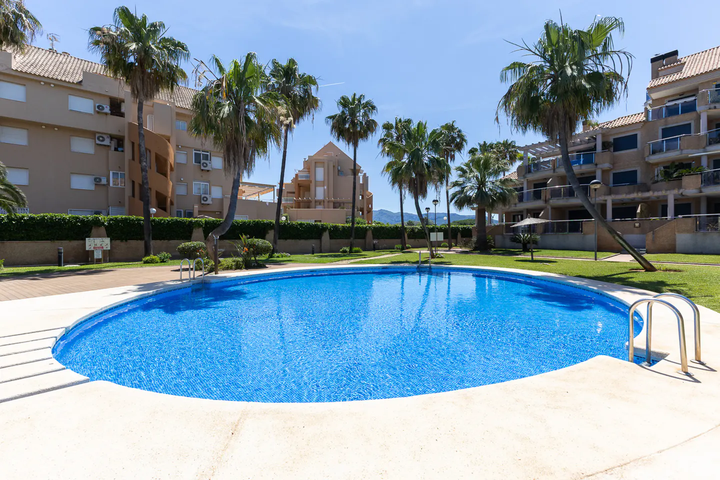 A round, blue swimming pool is surrounded by palm trees and apartment buildings on a sunny day.
