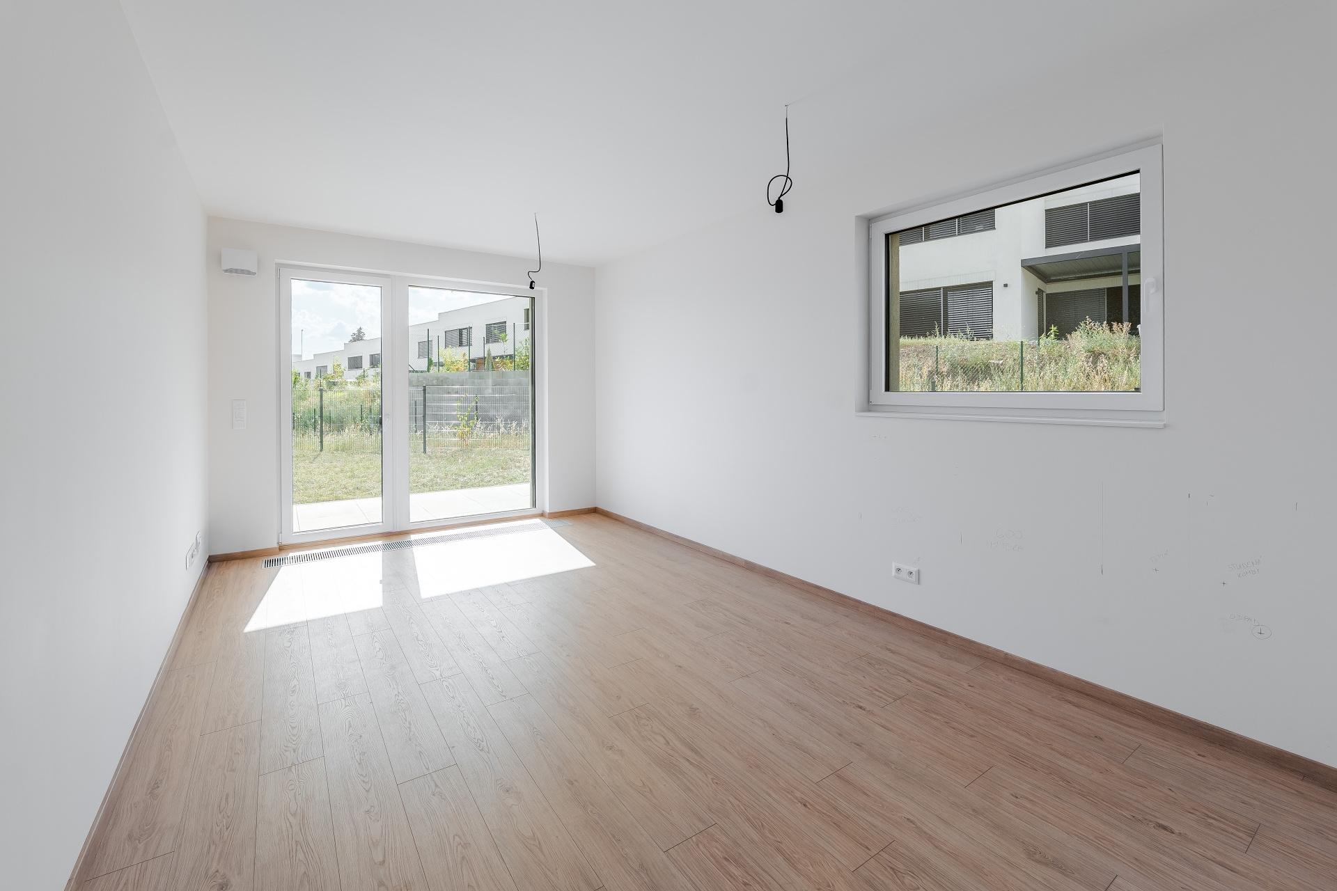 Empty room with white walls, light wood flooring, a glass door to a yard, and a window overlooking modern houses. [AI-generated text]