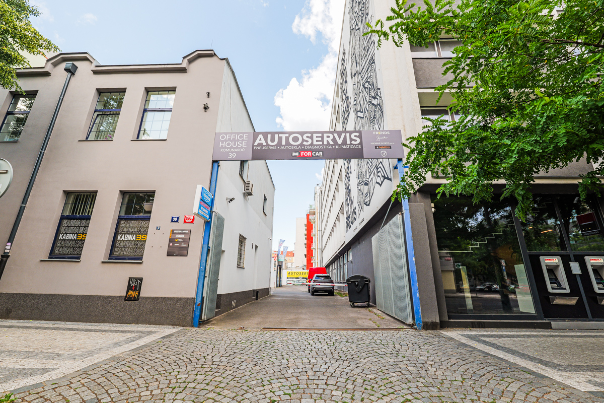 A narrow driveway between two buildings with an "Autoservis" sign overhead and a cobblestone street in the foreground.