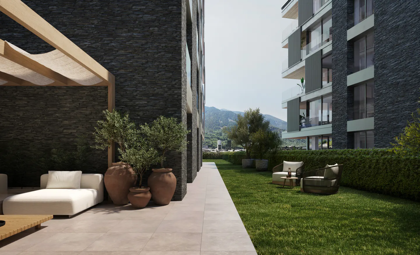 Modern apartment patio with white lounge furniture, potted olive trees, and a grassy lawn against a dark stone building.