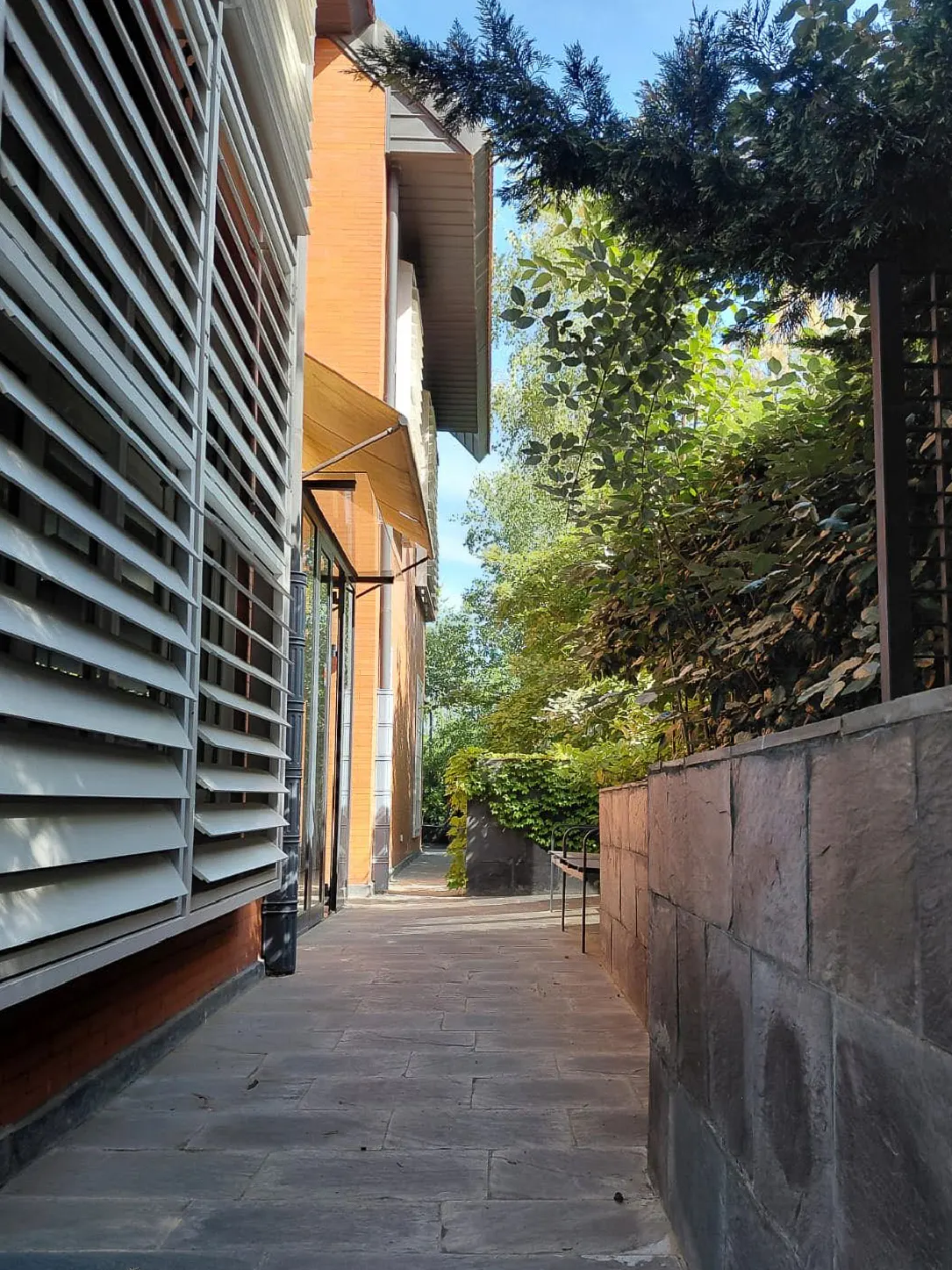 Exterior view of a building with white louvered windows, an orange brick facade, and a stone walkway leading to lush greenery.