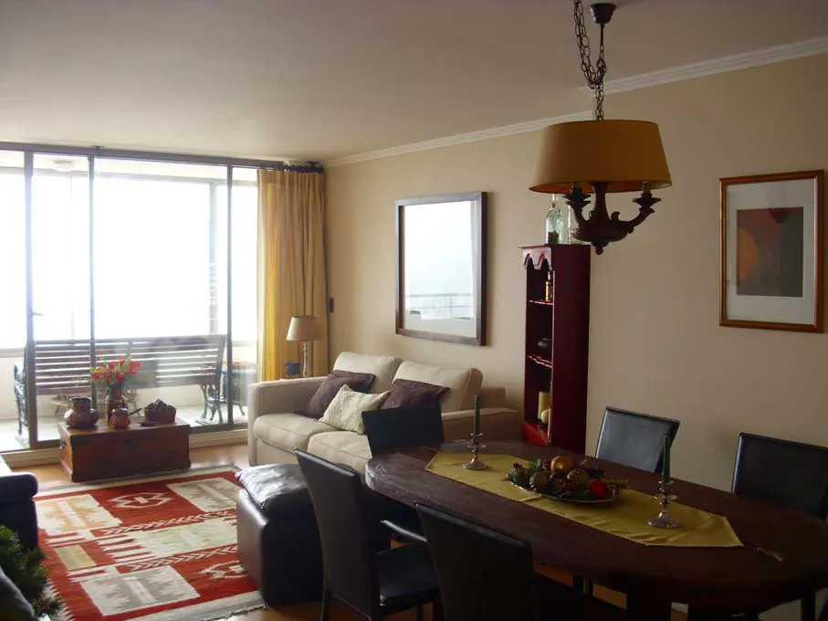 Living room and dining area with beige walls, a tan sofa, and a dark wood dining table with black chairs. A balcony is visible through sliding glass doors.