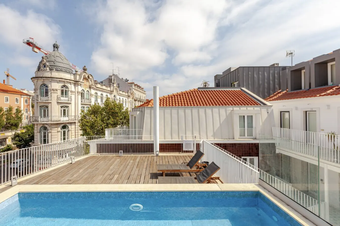 Rooftop pool with two lounge chairs on a wooden deck. White buildings and a blue sky with clouds in the background.