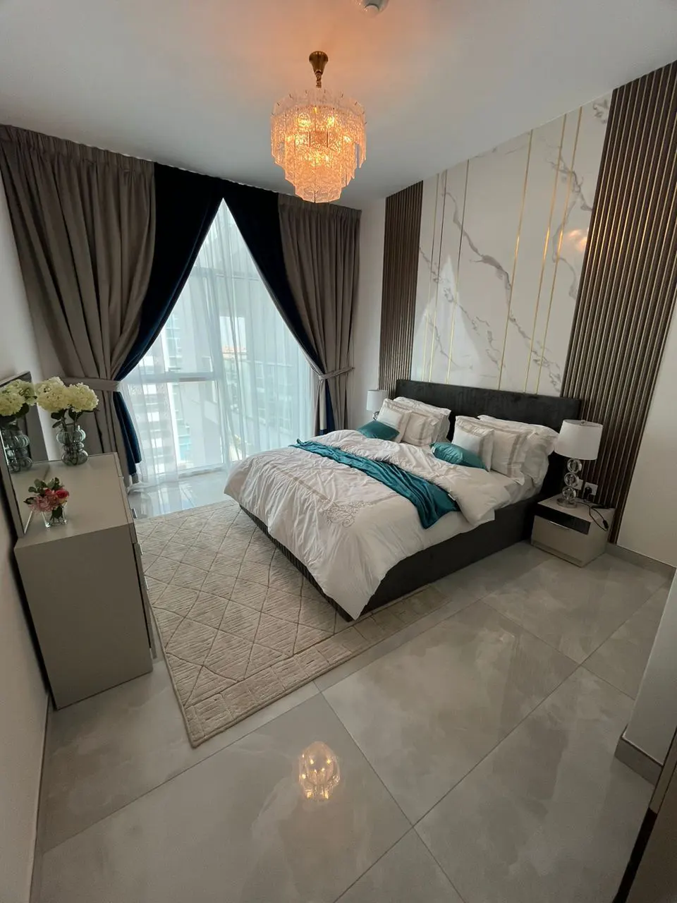 Bedroom with a king bed, white bedding, teal throw, and a crystal chandelier. Marble accent wall with gold trim and wood slats. Gray curtains frame a large window.