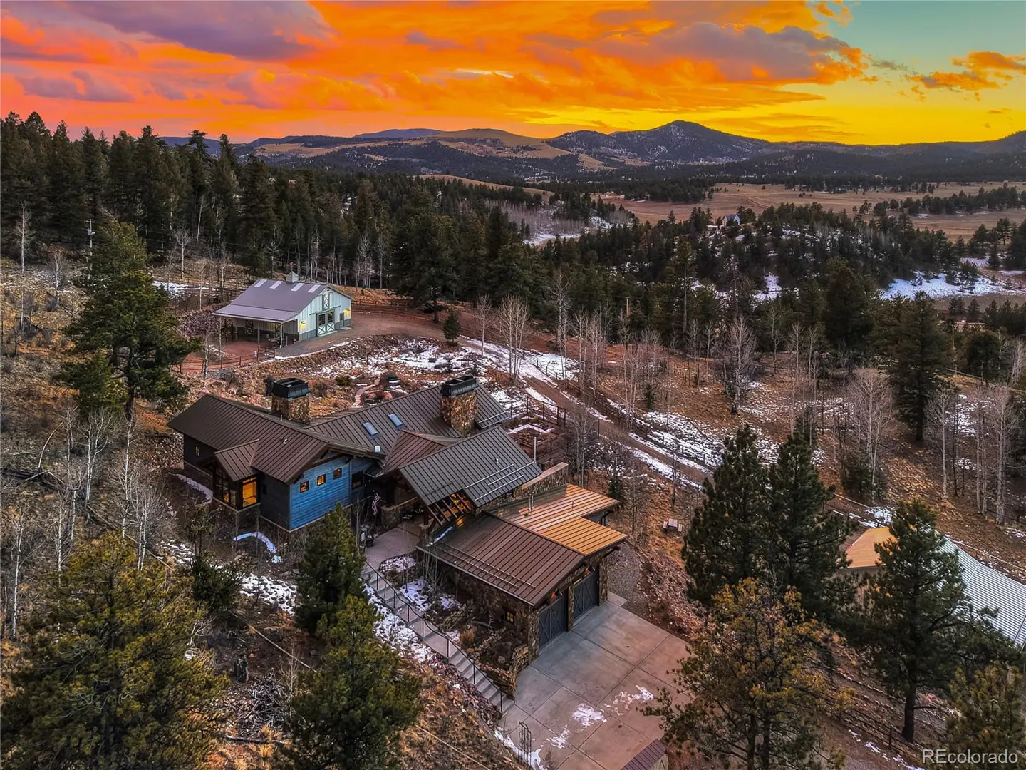 Aerial view of a luxury home with a stone facade and metal roof, nestled in a wooded area with mountains and a vibrant sunset in the background.