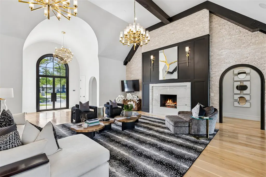 A bright living room with a white sofa, black chairs, and a fireplace. A black and white rug covers the wood floor. Chandeliers hang from the high ceiling.