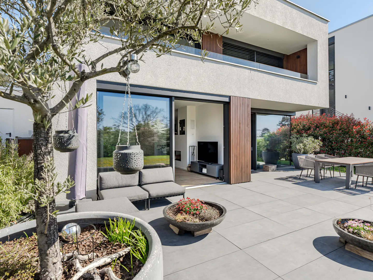 Modern home exterior with a gray patio, outdoor furniture, and potted plants. A tree with hanging lanterns adds charm.