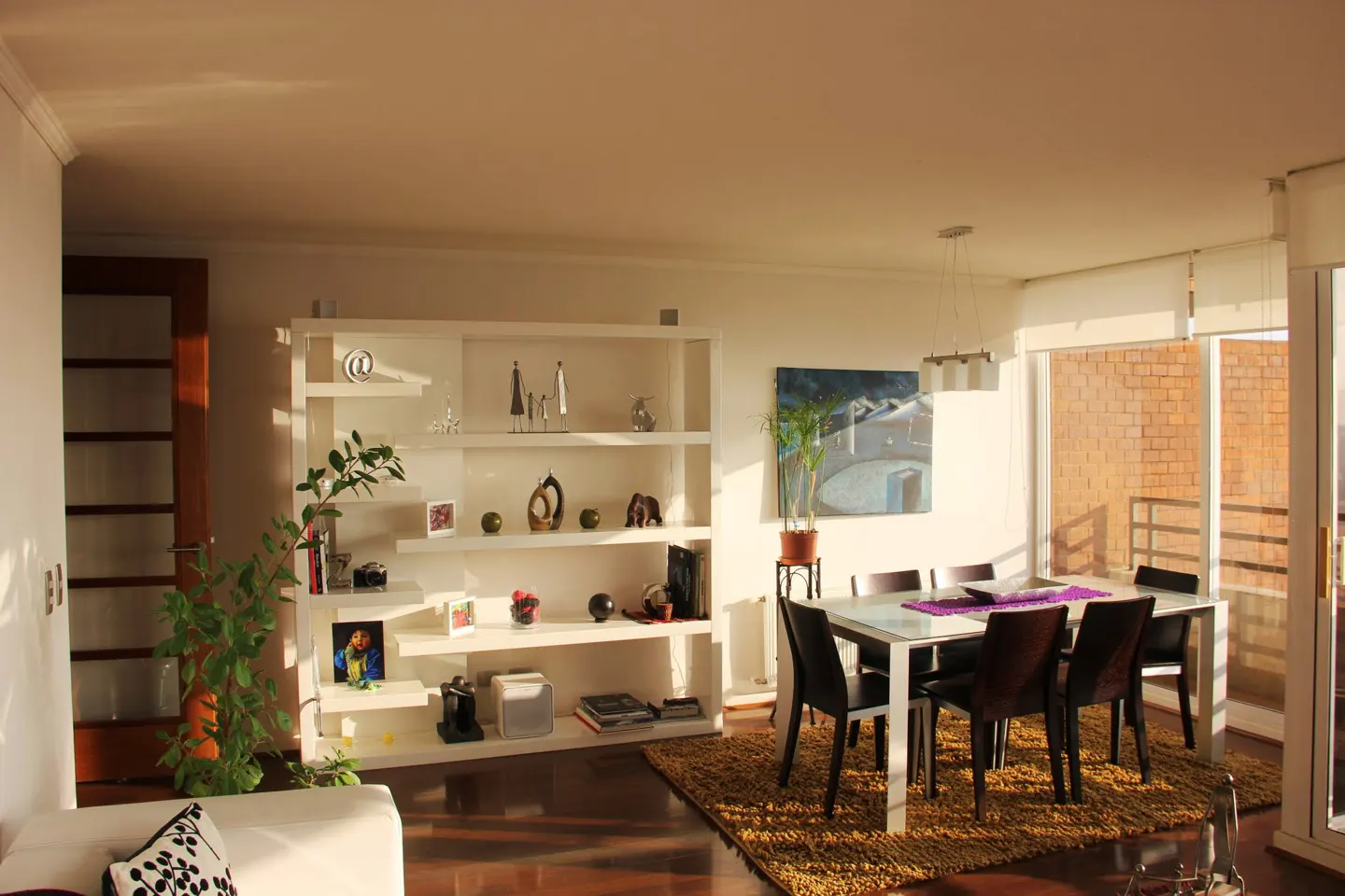 Bright living room with a white bookshelf, dining table with six chairs, and a large window with sunlight streaming in.