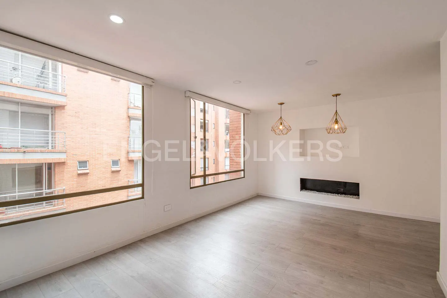 Bright, empty living room with gray wood floors, white walls, and two large windows showing a brick building. Two pendant lights hang above a modern fireplace.