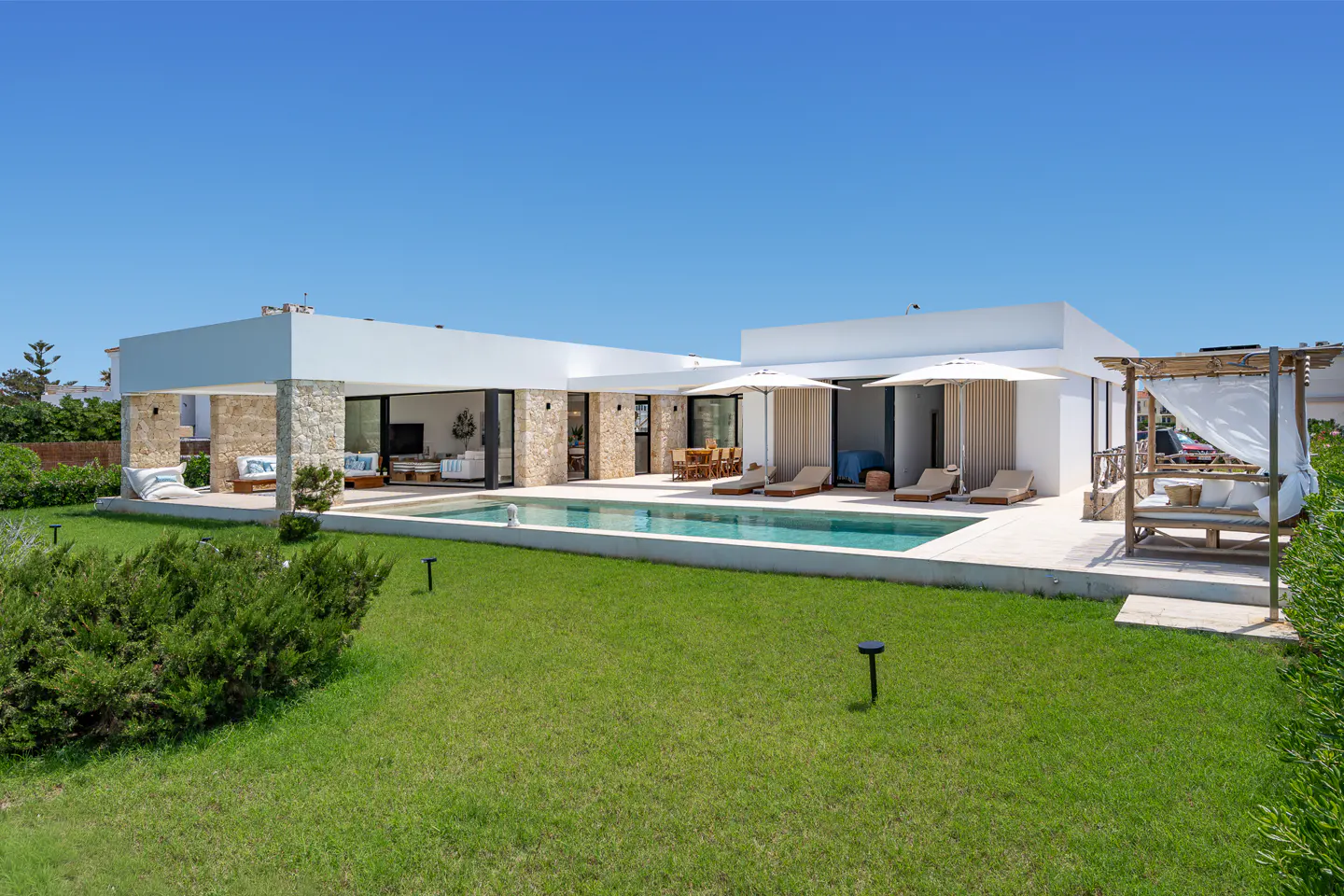 Modern white villa with a swimming pool, stone accents, and lounge chairs on a sunny lawn under a clear blue sky.