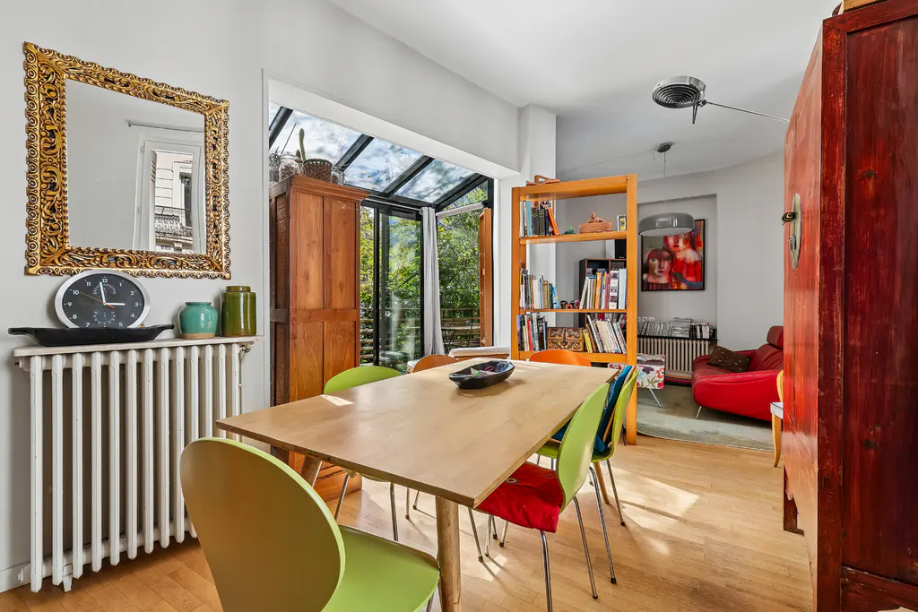 Bright dining room with a wooden table, colorful chairs, and a view to a garden through glass doors. A gold-framed mirror hangs above a radiator.