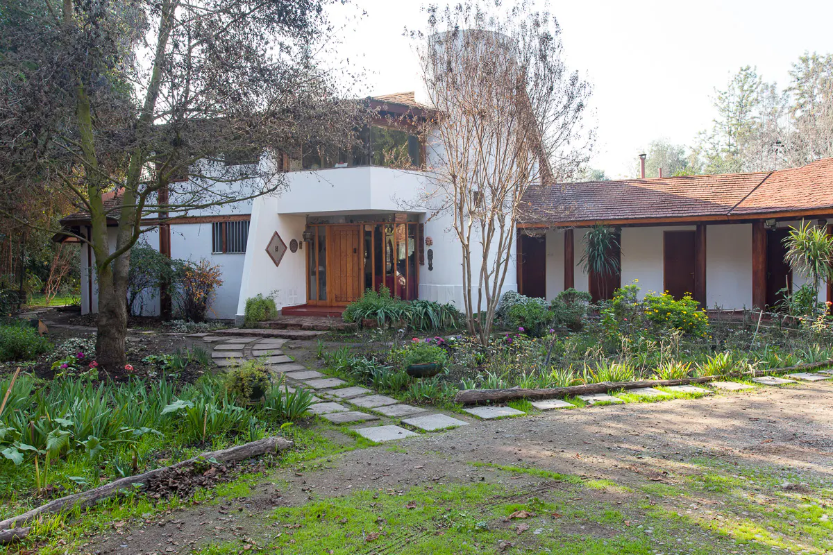 Exterior of a white house with a brown roof and wooden door, surrounded by a garden and trees.