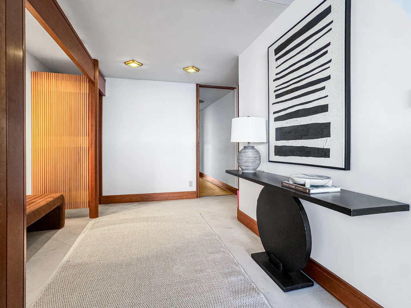Hallway with white walls, light carpet, and wood trim. A black console table with a lamp and art is on the right. A wood screen is on the left.