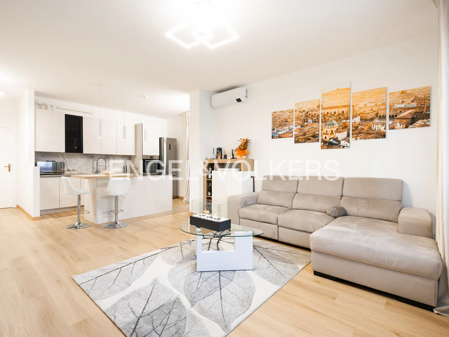 Bright, open-concept living space with a beige sectional sofa, leaf-patterned rug, and white kitchen with a breakfast bar.