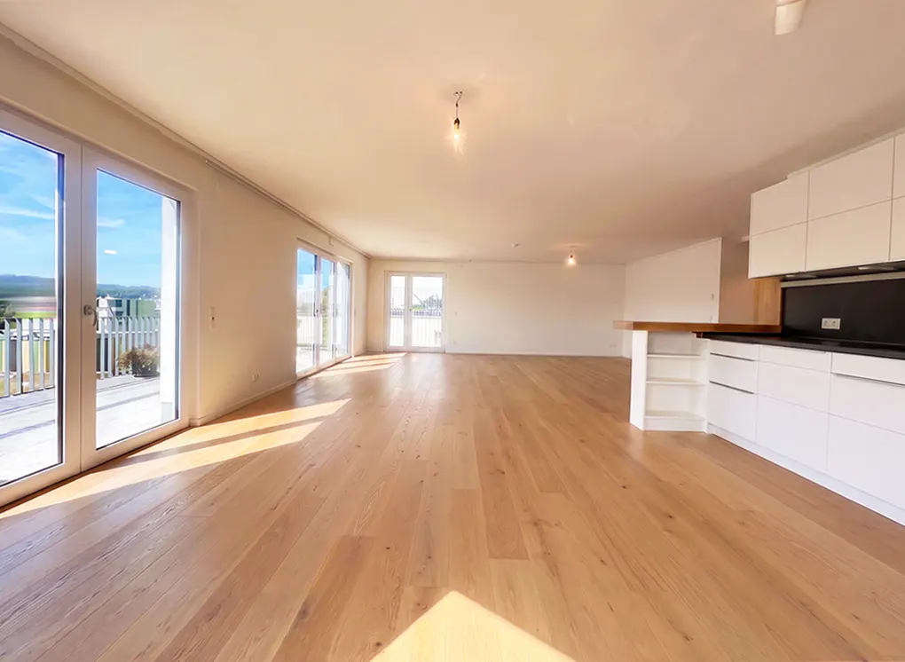 Spacious, sunlit open-plan living area with light wood floors, white kitchen cabinets, and glass doors leading to a balcony.