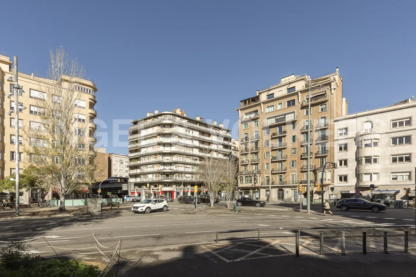 Cityscape view of a square with beige buildings, trees, cars, and pedestrians under a clear blue sky.