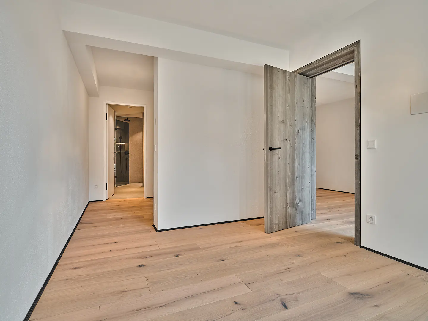 Bright room with light wood floors, white walls, and two open doorways. One doorway has a gray wood door ajar.