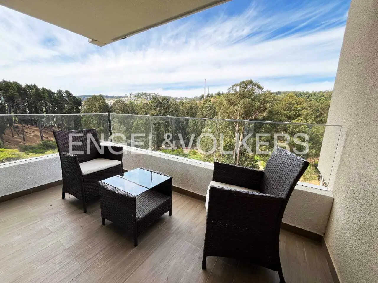 Balcony with two dark wicker chairs, a glass-topped table, and a view of trees and sky.