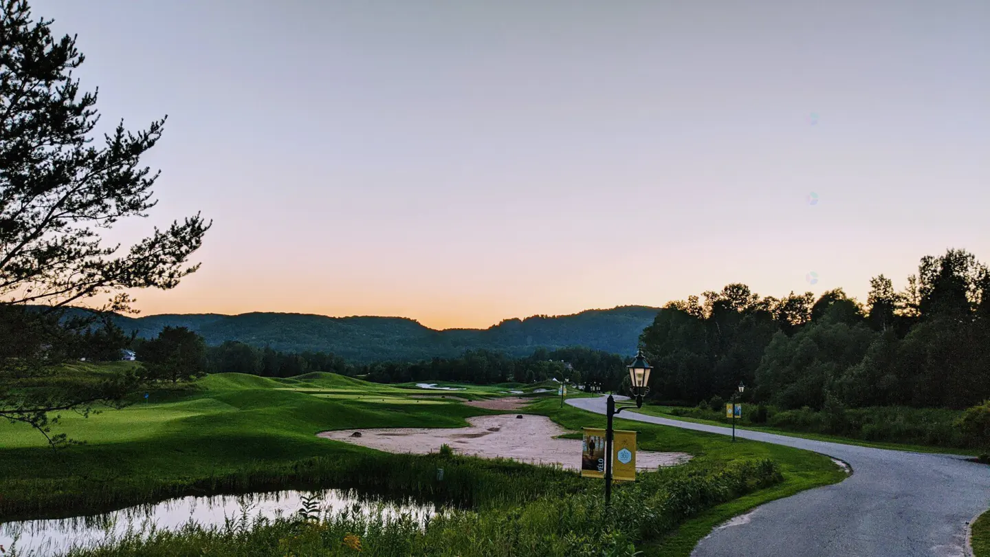 Scenic view of a golf course at sunset. Green hills, sand traps, and a winding path are visible. A pond and trees frame the foreground.
