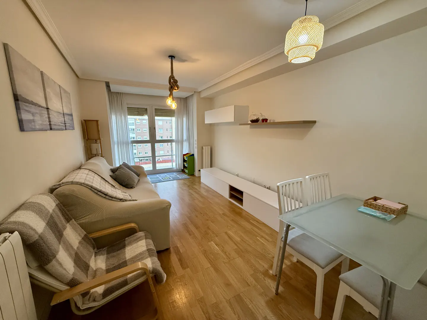 Bright living room with wood floors, a beige sofa, a glass dining table with white chairs, and modern lighting fixtures.